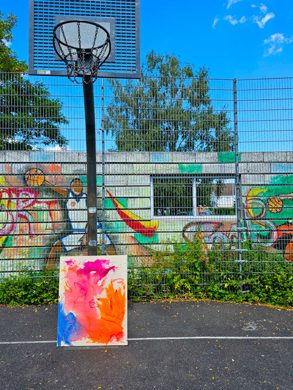 Bunte Kunst auf Basketballplatz vor graffiti-bedeckter Wand und blauem Himmel.