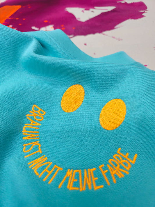Nahaufnahme des türkisen Sweaters mit neon oranger Stickerei eines Smileys und dem Schriftzug „Braun ist nicht meine Farbe“ – Detailansicht der hochwertigen Stickqualität.
