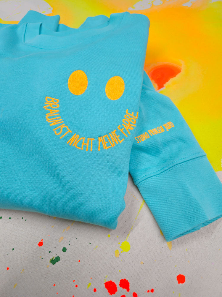 Gefalteter Sweater mit Smiley-Stickerei auf leuchtend gelbem Hintergrund – farbenfrohes Statement für Vielfalt und Haltung.