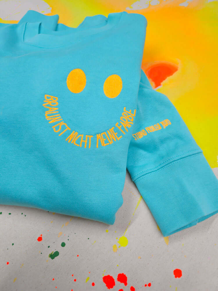Gefalteter Sweater mit Smiley-Stickerei auf leuchtend gelbem Hintergrund – farbenfrohes Statement für Vielfalt und Haltung.