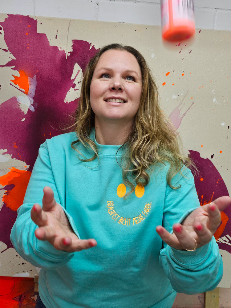 Künstlerin Studio Froilein Juno x Alexandra Wolf trägt den Statement-Sweater im Atelier, lachend mit neonfarbenem Farbtopf – kreativer Ausdruck in nachhaltiger Mode.