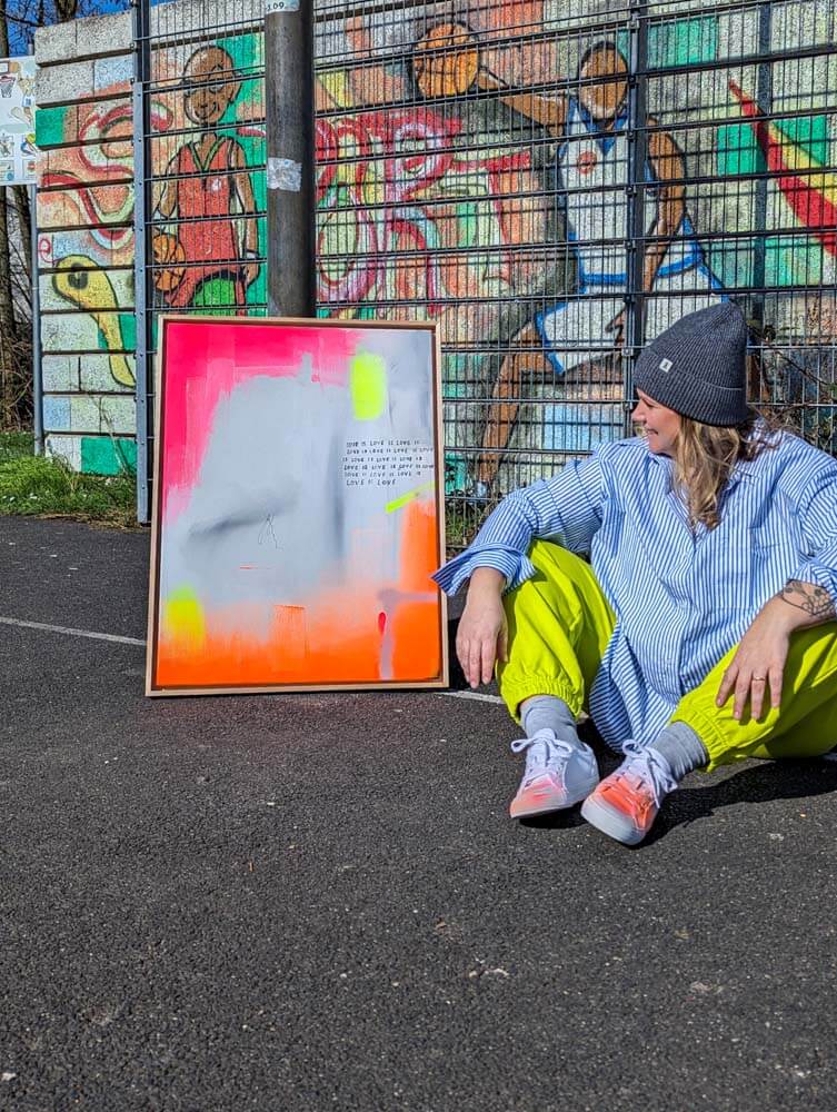 Person seated next to colorful mixed media artwork on pavement, urban graffiti backdrop, casual style.