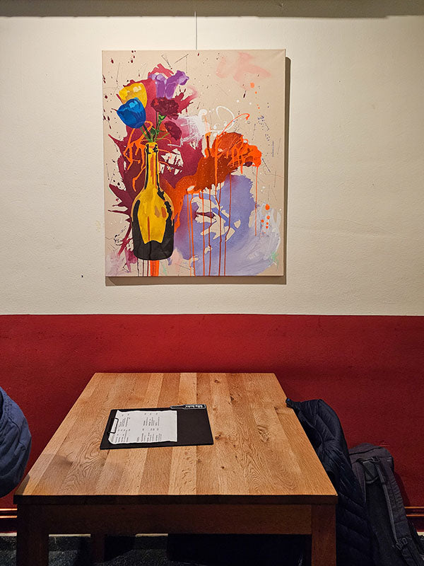 Gemälde „Flowers“ über einem Holztisch in einer Bonner Kneipe, abstrakte Blütenfarben und gelbe Flasche vor heller Wand.