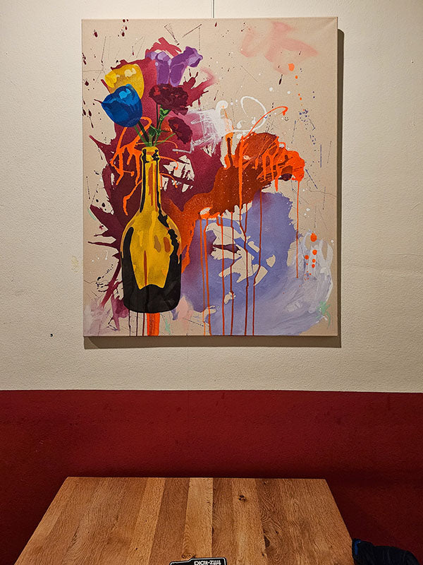 Abstraktes Kunstwerk „Flowers“ von Studio Froilein Juno und Baggadeluxe, hängend über Tischkante, kräftige Farbspritzer und florale Formen.