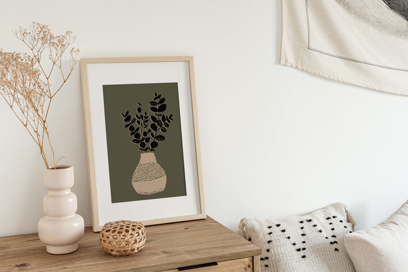 In einem Boho Schlafzimmer auf einer Kommode zeigt ein gerahmter Kunstdruck in offener Edition eine Vase mit ein paar Zweigen darin auf grünem Grund