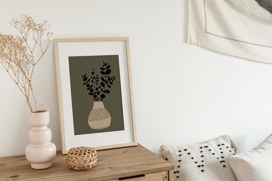 In einem Boho Schlafzimmer auf einer Kommode zeigt ein gerahmter Kunstdruck in offener Edition eine Vase mit ein paar Zweigen darin auf grünem Grund